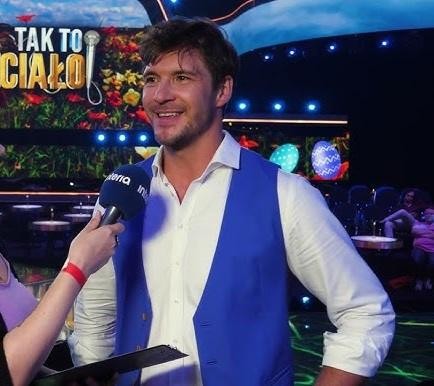 Mikołaj Roznerski na planie filmowym, w kostiumie, w trakcie pracy, z ekipą w tle.