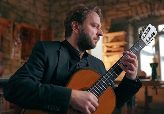 Marcin Dylla, gitarzysta klasyczny, grający na scenie w sali koncertowej, skupiony na gitarze, z publicznością w tle.
