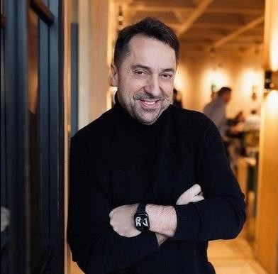 Marcin Miller pozujący obok czerwonego Mercedesa Cabrio i luksusowego Lexusa, uśmiechnięty i pełen optymizmu, w tle subtelny krajobraz.