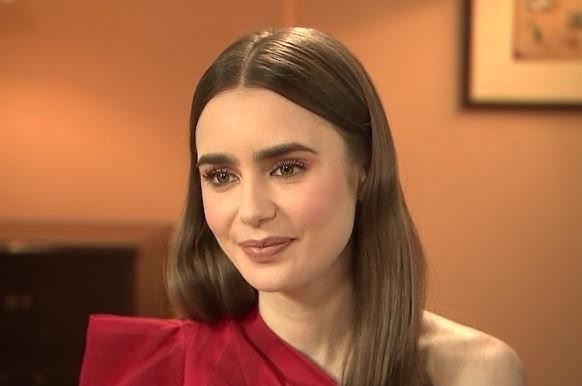 Lily Collins na planie filmowym, śmiejąca się, w stroju nawiązującym do postaci, z elementami scenografii w tle.