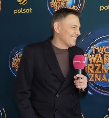 Krzysztof Ibisz prowadzący teleturniej w studiu telewizyjnym, z uśmiechem i gestykulacją, w tle kolorowe elementy scenografii.